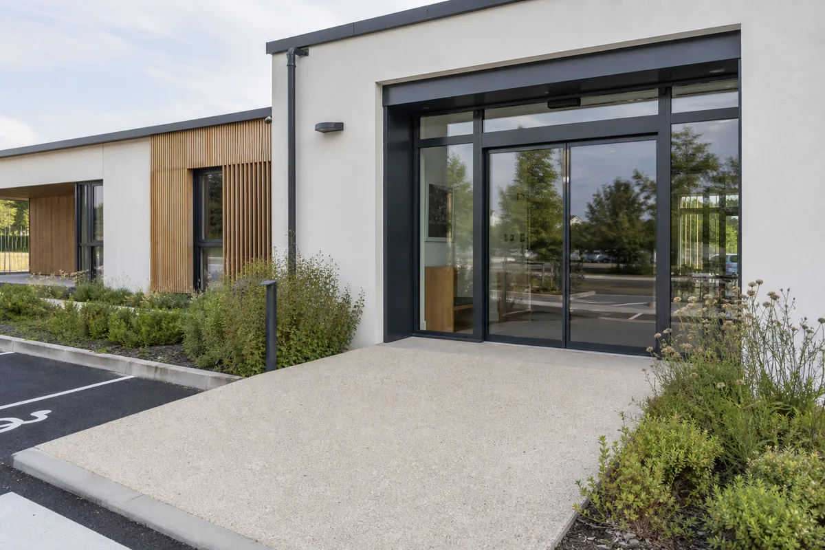 Entrée accessible d&rsquo;un bâtiment recevant du public avec rampe douce, seuil de plain-pied et menuiserie extérieure soignée, cheminement relié au parking